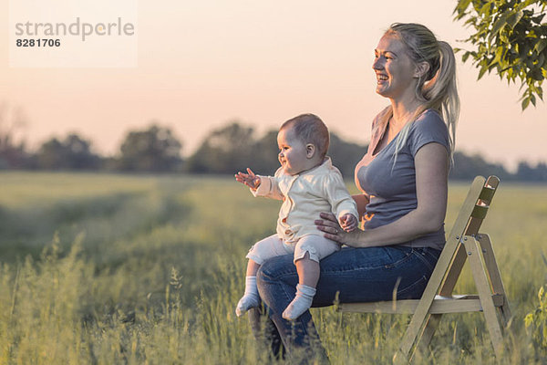 Eine lachende Frau hält ihr Baby  während sie auf einem Stuhl auf einem Feld sitzt.