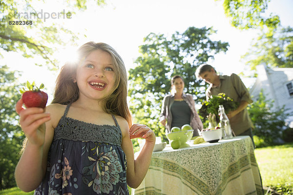 Auf der Farm. Kinder und Erwachsene zusammen. Ein junges Mädchen hält eine große frische  biologisch erzeugte Erdbeere in der Hand. Zwei Erwachsene neben einem runden Tisch.