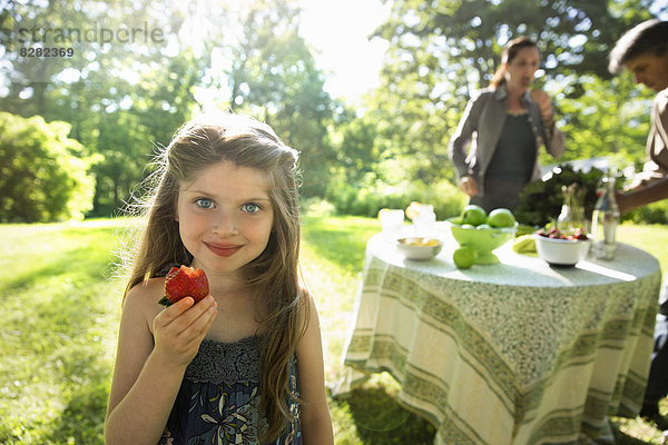 Ein junges Mädchen mit einer großen frischen  biologisch produzierten Erdbeere. Zwei Erwachsene neben einem runden Tisch.