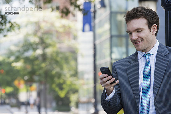 Sommer. Ein junger Mann in einem grauen Anzug und blauer Krawatte. Mit einem Smartphone.