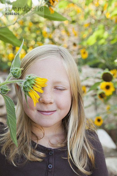 Ein Kind steht in einem Blumengarten. Ein Mädchen mit geschlossenen Augen und einer Sonnenblume vor dem Auge.