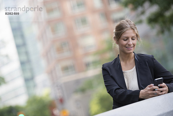 Eine junge blonde Geschäftsfrau auf einer Straße in New York City. Trägt eine schwarze Jacke. Benutzt ein Smartphone.