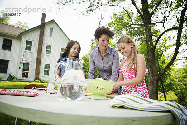 Eine Sommer-Familienzusammenkunft auf einem Bauernhof. Eine Frau und zwei Kinder stehen draußen an einem Tisch und decken den Tisch. Sie machen Limonade.