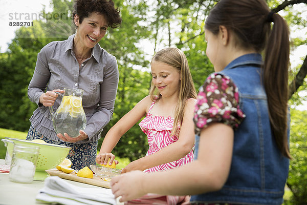 Eine Sommer-Familienzusammenkunft auf einem Bauernhof. Eine Frau und zwei Kinder stehen draußen an einem Tisch und decken den Tisch. Sie machen Limonade.