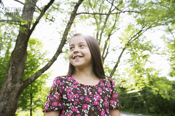 Ein junges Mädchen in einem gemusterten Sommerkleid  unter dem Schatten von Bäumen in einem Bauerngarten.