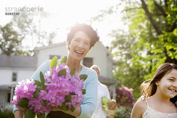 Familienfeier. Eine Frau  die einen großen Strauß Rhododendronblumen trägt und breit lächelt.