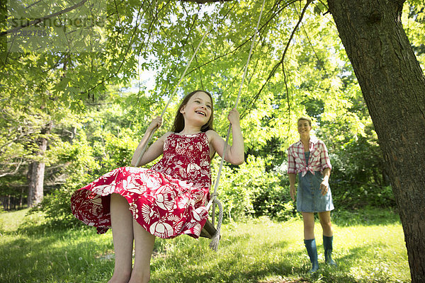 Sommer. Ein Mädchen in einem Sonnenkleid auf einer Schaukel unter einem blattreichen Baum. Eine Frau  die hinter ihr steht.