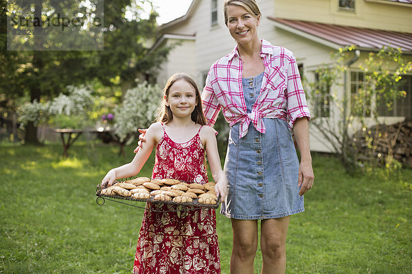 Hausgemachte Kekse backen. Ein junges Mädchen hält ein Tablett mit frisch gebackenen Keksen in der Hand  daneben eine erwachsene Frau.