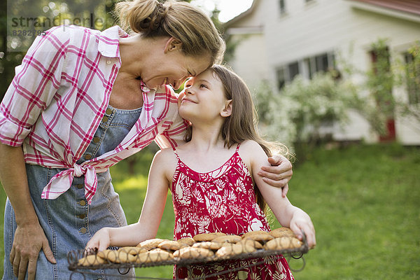 Hausgemachte Kekse backen. Ein junges Mädchen hält ein Tablett mit frisch gebackenen Keksen in der Hand  und eine erwachsene Frau beugt sich vor  um sie zu loben.