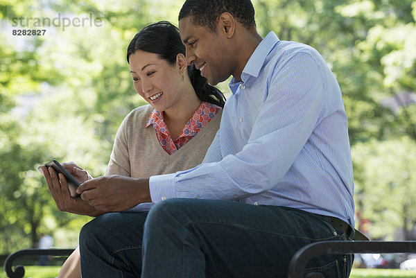 Sommer. Geschäftsleute. Ein Mann und eine Frau sitzen auf einer Bank und benutzen ein Smartphone.
