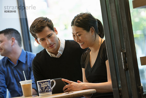 Eine Gruppe von Menschen  die um einen Tisch in einem Coffee Shop sitzen. Sie schauen auf den Bildschirm eines digitalen Tabletts. Zwei Männer und eine Frau.
