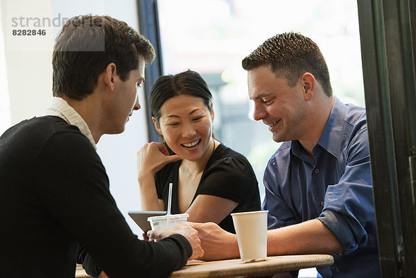 Eine Gruppe von Menschen  die um einen Tisch in einem Coffee Shop sitzen. Sie schauen auf den Bildschirm eines digitalen Tabletts. Zwei Männer und eine Frau.