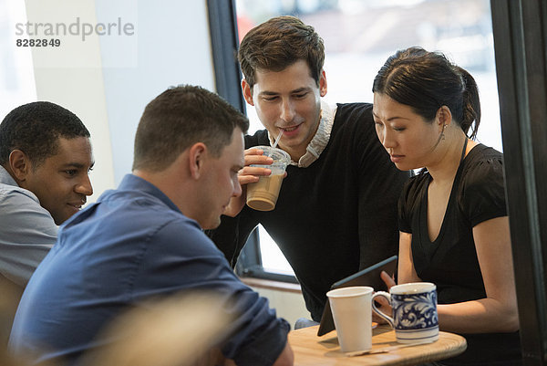Eine Gruppe von Menschen  die um einen Tisch in einem Coffee Shop sitzen. Sie schauen auf den Bildschirm eines digitalen Tabletts. Drei Männer und eine Frau.