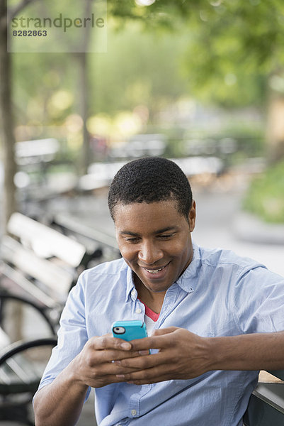 Sommer in der Stadt. Ein Mann sitzt auf einer Bank und benutzt ein Smartphone.