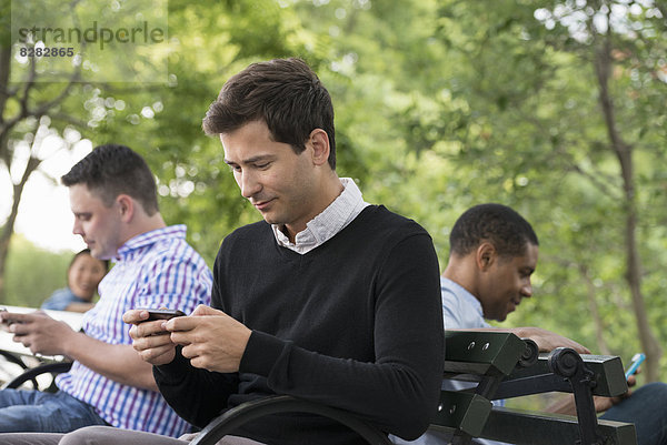 Sommer in der Stadt. Eine Frau und drei Männer sitzen im Park  jeder mit seinem eigenen Telefon oder mit einem Tablet.