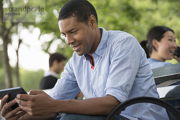 Sommer. Ein Mann sitzt auf einer Bank und benutzt ein digitales Tablett.
