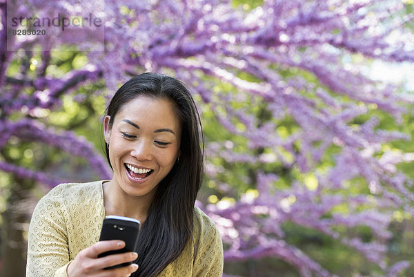 Eine Frau sitzt in einem Park  schaut auf ihr Smartphone und lacht.