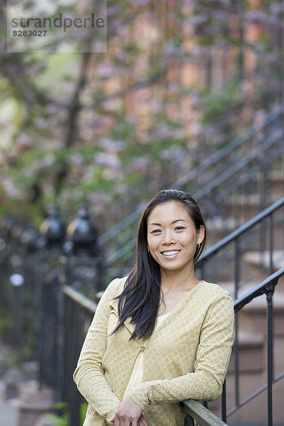 Eine junge Frau mit langen schwarzen Haaren  die sich am Fuße einer Treppe an ein Geländer gelehnt hat.