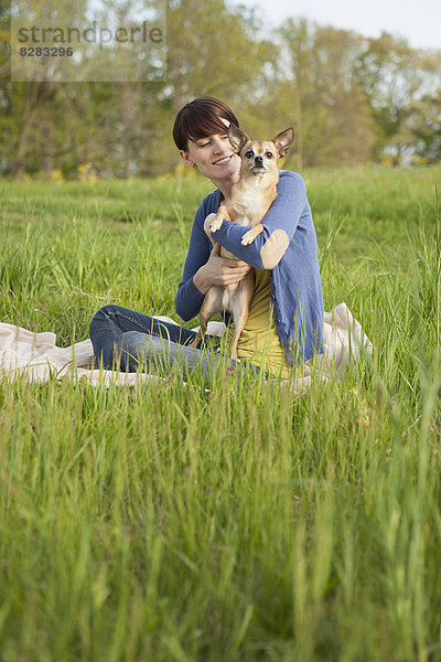 Eine junge Frau sitzt auf einem Feld  auf einer Decke und hält einen kleinen Chihuahua-Hund.