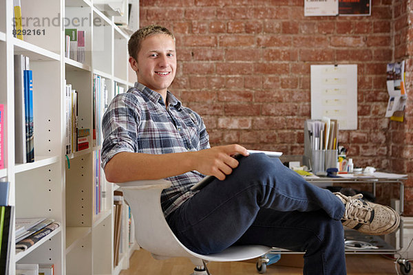 Porträt eines jungen Mannes  der auf einem Stuhl sitzt und ein digitales Tablett benutzt.