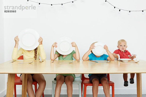 Vier Kinder am Tisch sitzend  drei Gesichter mit Tellern bedeckt