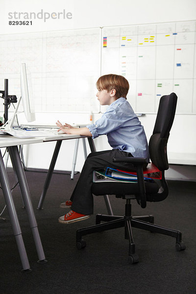 Junge sitzt auf Ringbüchern auf Bürostuhl mit Computer