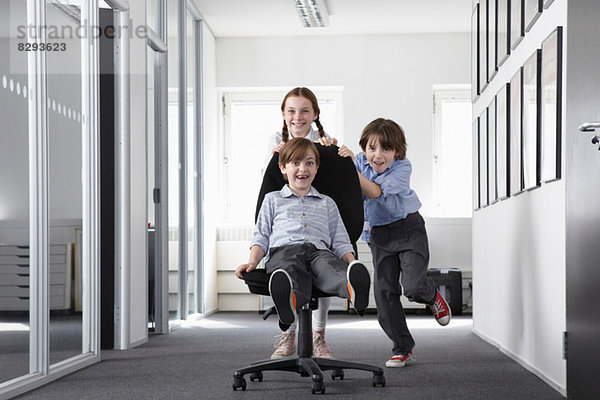 Drei Kinder spielen im Büroflur auf Bürostuhl