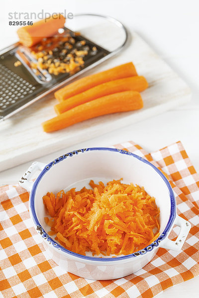 Geriebene Karotten in einer Schüssel  Karotten und Reibe im Hintergrund