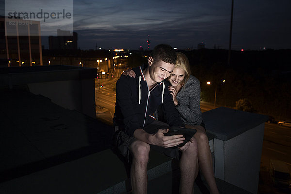 Junges Paar mit Tablett-PC auf dem Flachdach bei Nacht