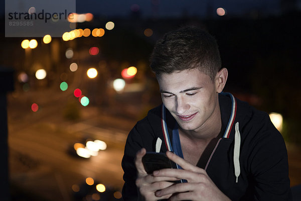 Young man with smartphone sitting on flat roof by night