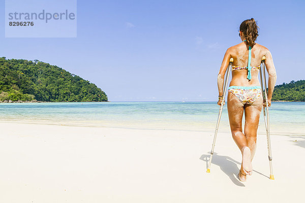 Thailand  Insel Koh Surin  Frau mit Krücken  die am weißen Sandstrand spazieren geht.