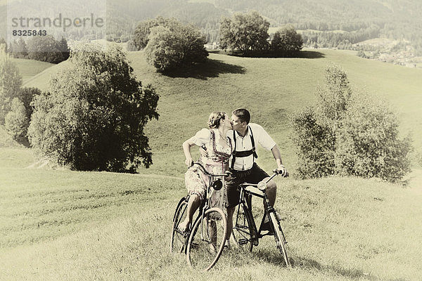 Mann in Lederhose und Frau in Dirndl küssen sich auf alten Fahrrädern in der Natur
