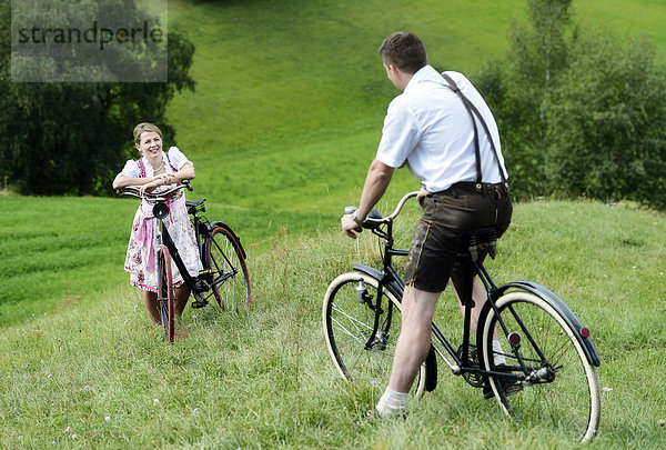 Mann in Lederhose und Frau in Dirndl auf alten Fahrrädern in der Natur