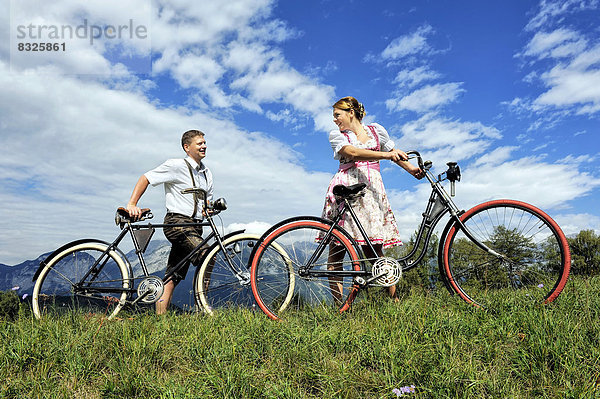 Mann in Lederhose und Frau in Dirndl auf alten Fahrrädern in der Natur