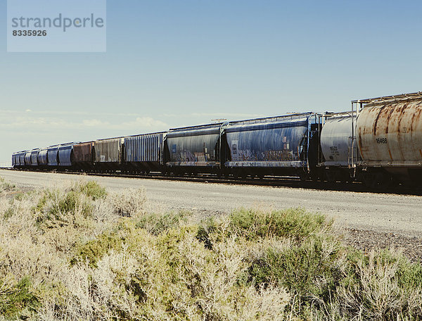 Reihe von Container-Güterwagen eines Güterzuges  der die Ebene bei Wendover durchquert. Gestrüppte Salbeibüsche  die an den Gleisen wachsen.