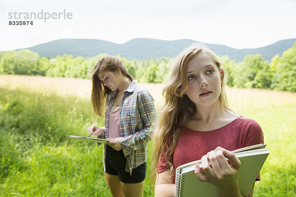 Zwei junge Mädchen sitzen draußen auf dem Rasen  mit Skizzenblöcken und Bleistiften.