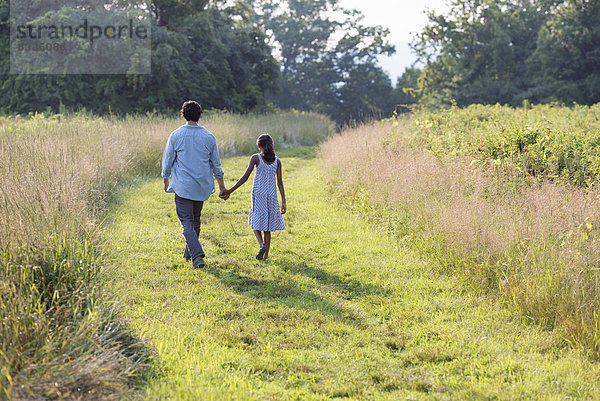 Ein Mann und ein junges Mädchen gehen einen gemähten Weg im langen Gras entlang und halten sich an den Händen.