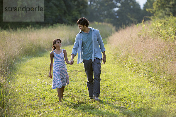 Ein Mann und ein junges Mädchen gehen einen gemähten Weg im langen Gras entlang und halten sich an den Händen.