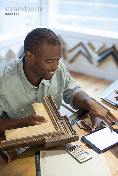 Ein junger Mann an seiner Werkbank in einem Bildereinrahmungsstudio. Umgeben von Mustern. Mit einem digitalen Tablett.