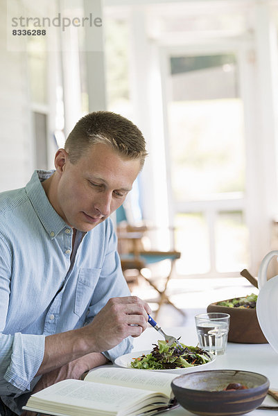 Ein Mann sitzt an einem Cafétisch  isst einen Salat und liest ein Buch.