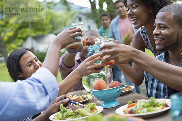 Eine Gruppe von Erwachsenen und Jugendlichen bei einer Mahlzeit im Garten eines Bauernhauses. Vorbei an Tellern und Gläsern.