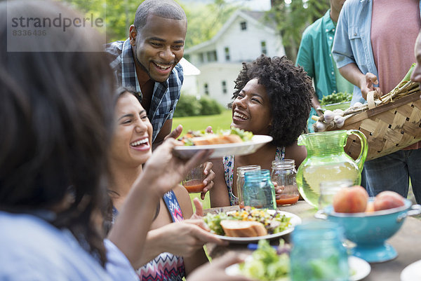 Eine Gruppe von Erwachsenen und Jugendlichen bei einer Mahlzeit im Garten eines Bauernhauses. Vorbei an Tellern und Gläsern.