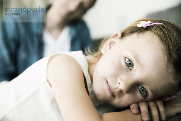 Nachdenkliches kleines Mädchen auf dem Sofa liegend  ihr Vater im Hintergrund stehend