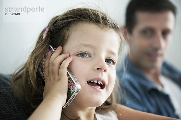 Porträt eines kleinen Mädchens  das mit dem Handy telefoniert und von seinem Vater beobachtet wird.
