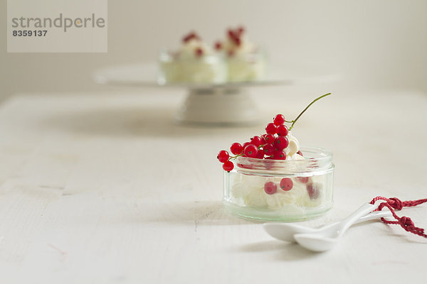 Joghurt-Sahne-Dessert mit roten Johannisbeeren im Glas  zwei weißen Löffeln und einem Kuchenstand mit Dessertgläsern.