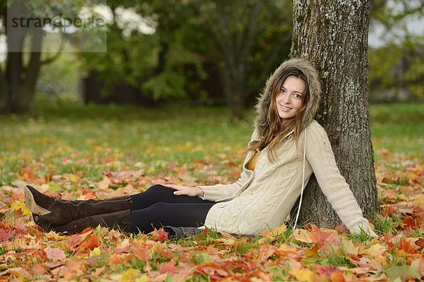 Lächelnde junge Frau sitzt unter einem Baum im Herbst