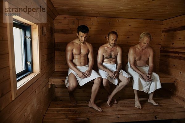 Drei Männer sitzen in der Sauna mit gebeugten Köpfen.