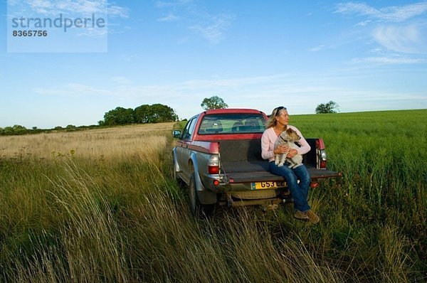 Frau sitzt auf dem Rücksitz eines Pick-up auf einem Feld und hält einen Hund.