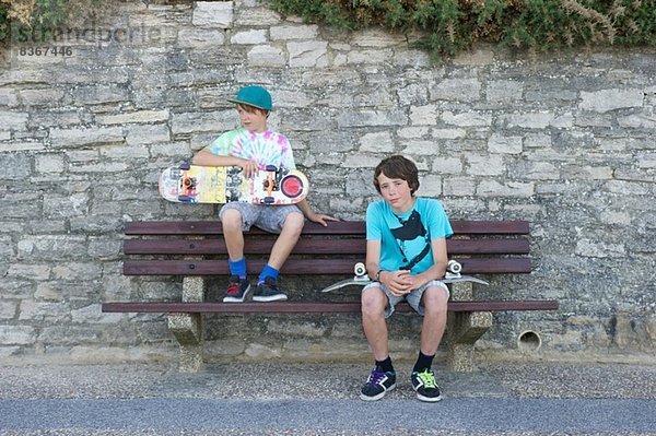 Porträt von zwei Jungen  die auf einer Bank sitzen und Skateboards halten.