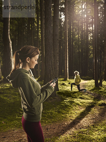 Frau steht im Wald und schaut auf ihr Smartphone  hinten macht eine andere Frau Dehnübungen  Tirol  Austria